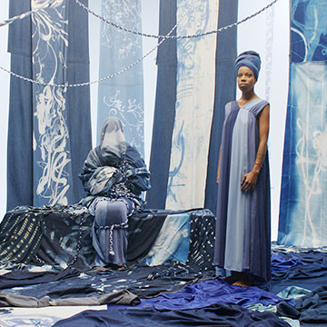 a woman stands next to a shrouded seated figure. They are surrounded by blue/white/gray fabrics all around